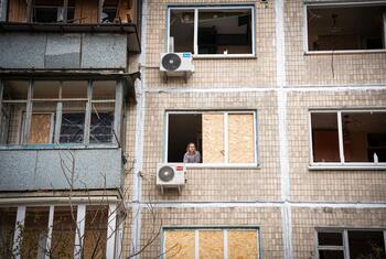 A woman looks out from a window of an apartment building with damaged windows and boarded-up panes in Kyiv, Ukraine.