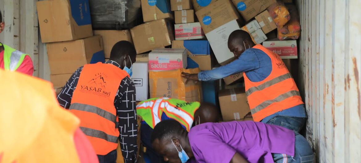 Des travailleurs humanitaires en gilets de sécurité orange chargent des caisses de fournitures médicales dans un conteneur d'expédition au sein d'un centre d'urgence de l'OMS.