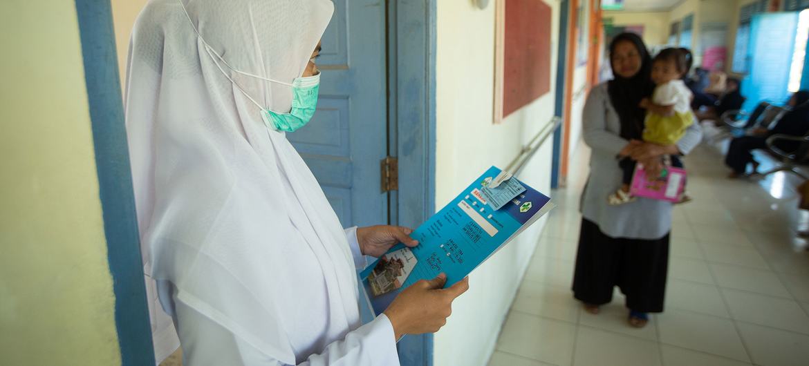 Uma enfermeira usando um hijab branco e uma máscara facial está no corredor de uma clínica, segurando uma pasta azul. Ao fundo, uma mãe segura uma criança pequena.