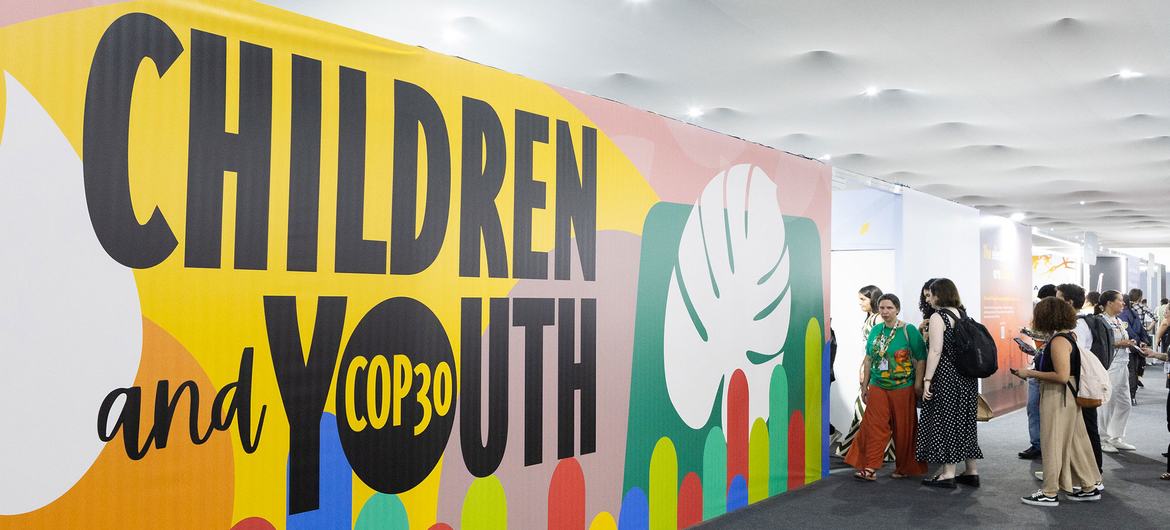 Uma faixa colorida com os dizeres "COP30 para Crianças e Jovens" é exibida em um evento, com participantes em pé nas proximidades.