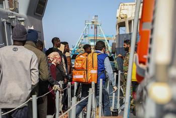 Un membre du personnel de l'OIM portant un sac à dos orange marqué "IOM RESCUE" aide des migrants à embarquer sur un navire en Libye, soulignant les efforts déployés par l'organisation pour prévenir la perte de vies sur la route centrale méditerranéenne.