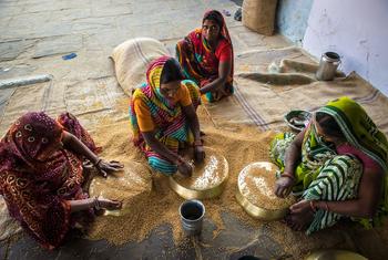 Four Indian women in colorful saris sit on the floor cleaning wheat grains in brass bowls.
