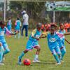 A group of young African children in blue soccer uniforms participate in a soccer training session on a grass field. One child in the foreground is dribbling a red and blue soccer ball, while others run and watch nearby.