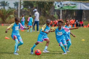 Un groupe de jeunes enfants africains, vêtus de maillots de football bleus, participent à un entraînement sur un terrain en herbe. Au premier plan, un enfant dribble un ballon rouge et bleu, tandis que d'autres courent et regardent à proximité.