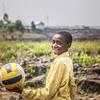 A smiling boy in a yellow shirt holds a volleyball at a UNICEF-supported child-friendly space in DR Congo.