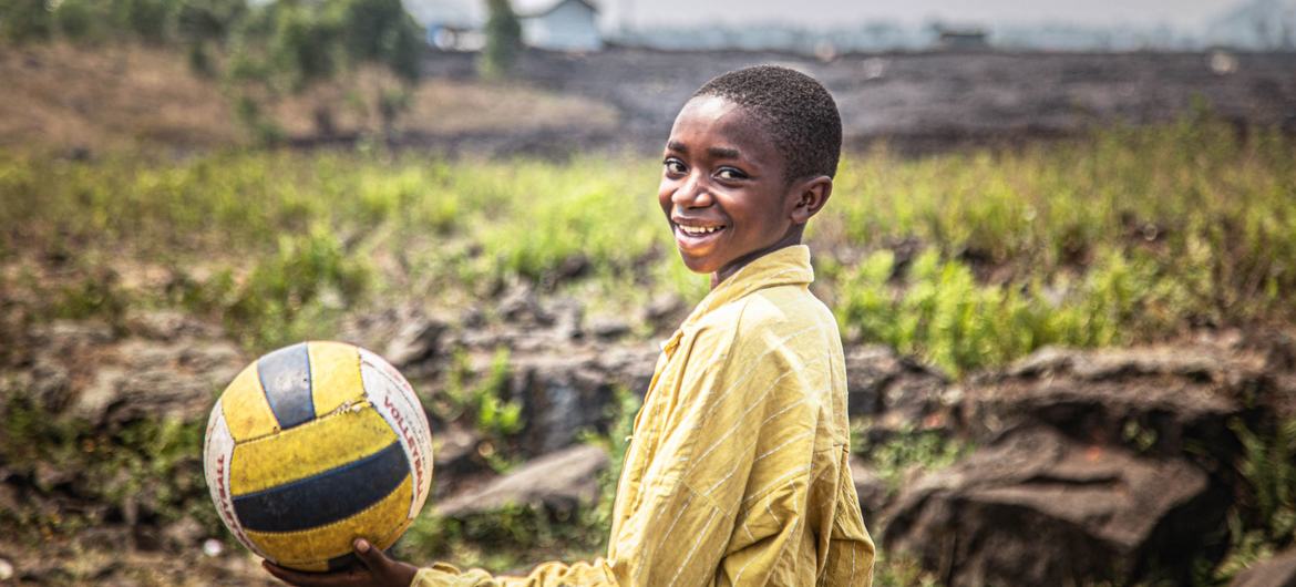 A smiling boy in a yellow shirt holds a volleyball at a UNICEF-supported child-friendly space in DR Congo.