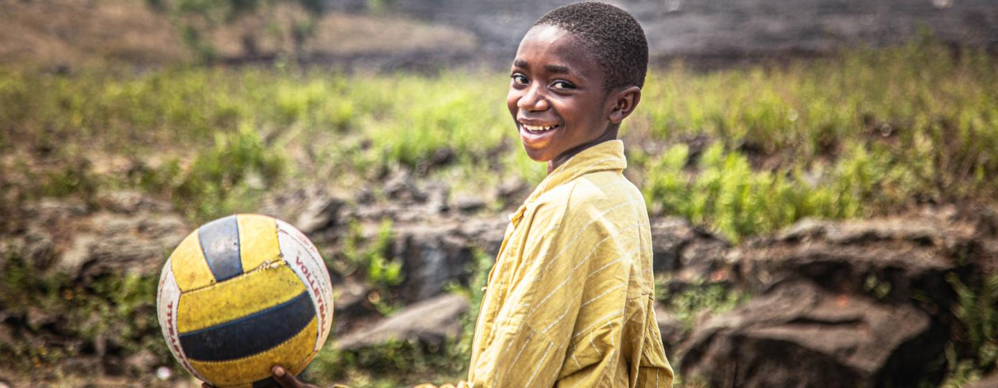 Un garçon souriant, vêtu d'un t-shirt jaune, tient un ballon de volley-ball dans un espace adapté aux enfants soutenu par l'UNICEF en République démocratique du Congo.