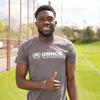 Alphonso Davies, akitabasamu na kuinua kidole chake huku amevaa T-shati ya UNHCR kwenye uwanja wa mpira wa miguu.
