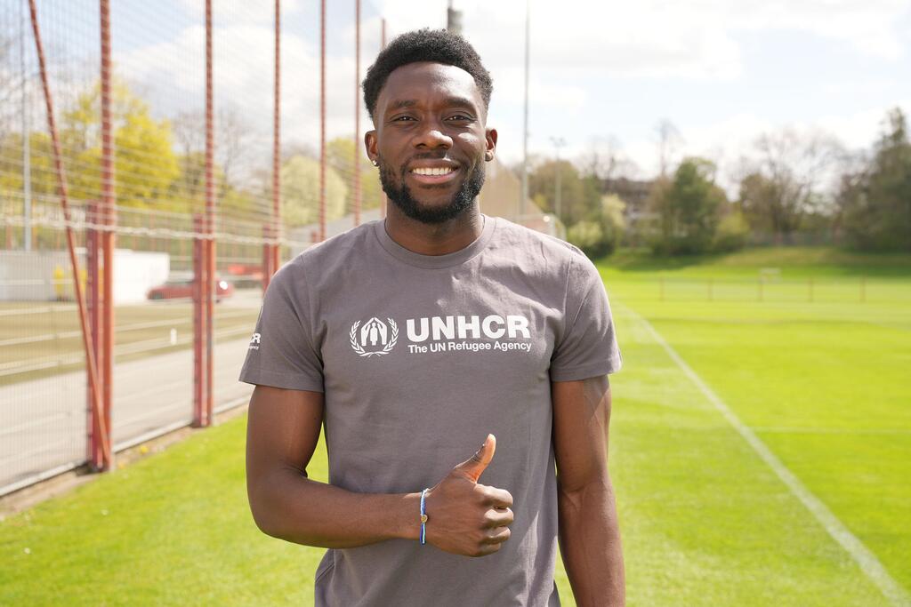 Alphonso Davies, souriant et faisant un pouce en l'air tout en portant un t-shirt du HCR sur un terrain de football.