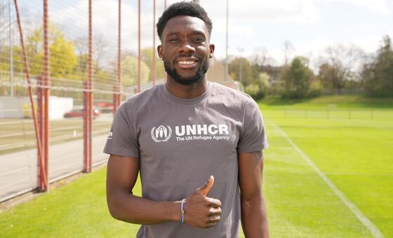 Alphonso Davies, akitabasamu na kuinua kidole chake huku amevaa T-shati ya UNHCR kwenye uwanja wa mpira wa miguu.