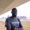 Mchezaji wa mpira wa miguu Antonio Rüdiger, amevaa T-shirt ya UNHCR, akitabasamu akiwa na mikono iliyounganishwa kwenye uwanja.