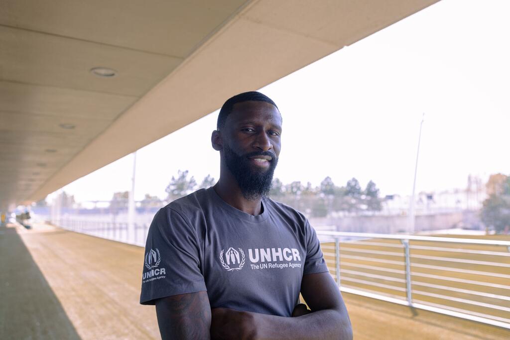 Mchezaji wa mpira wa miguu Antonio Rüdiger, amevaa T-shirt ya UNHCR, akitabasamu akiwa na mikono iliyounganishwa kwenye uwanja.