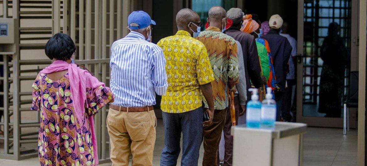 African adults wearing masks wait in a line at the entrance of a vaccination center in Kigali, Rwanda, near hand sanitizer dispensers and a metal turnstile.