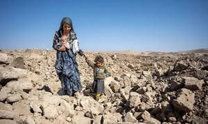A mother holding a baby and guiding a young child through a field of rubble in Afghanistan, likely after a flood, under a clear blue sky.