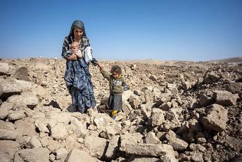 A mother holding a baby and guiding a young child through a field of rubble in Afghanistan, likely after a flood, under a clear blue sky.