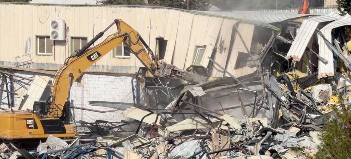 A yellow CAT excavator demolishes a building in East Jerusalem, UNRWA headquarters, creating a pile of rubble and debris.