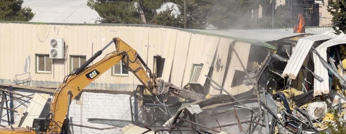 Une excavatrice CAT jaune démolit un bâtiment à Jérusalem-Est, siège de l'UNRWA, créant une pile de décombres et de débris.