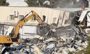 A yellow CAT excavator demolishes a building in East Jerusalem, UNRWA headquarters, creating a pile of rubble and debris.