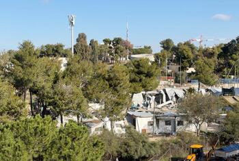 Une vue aérienne du siège de l'UNRWA à Jérusalem-Est en démolition, avec des bâtiments endommagés, des équipements de construction et des arbres environnants visibles.