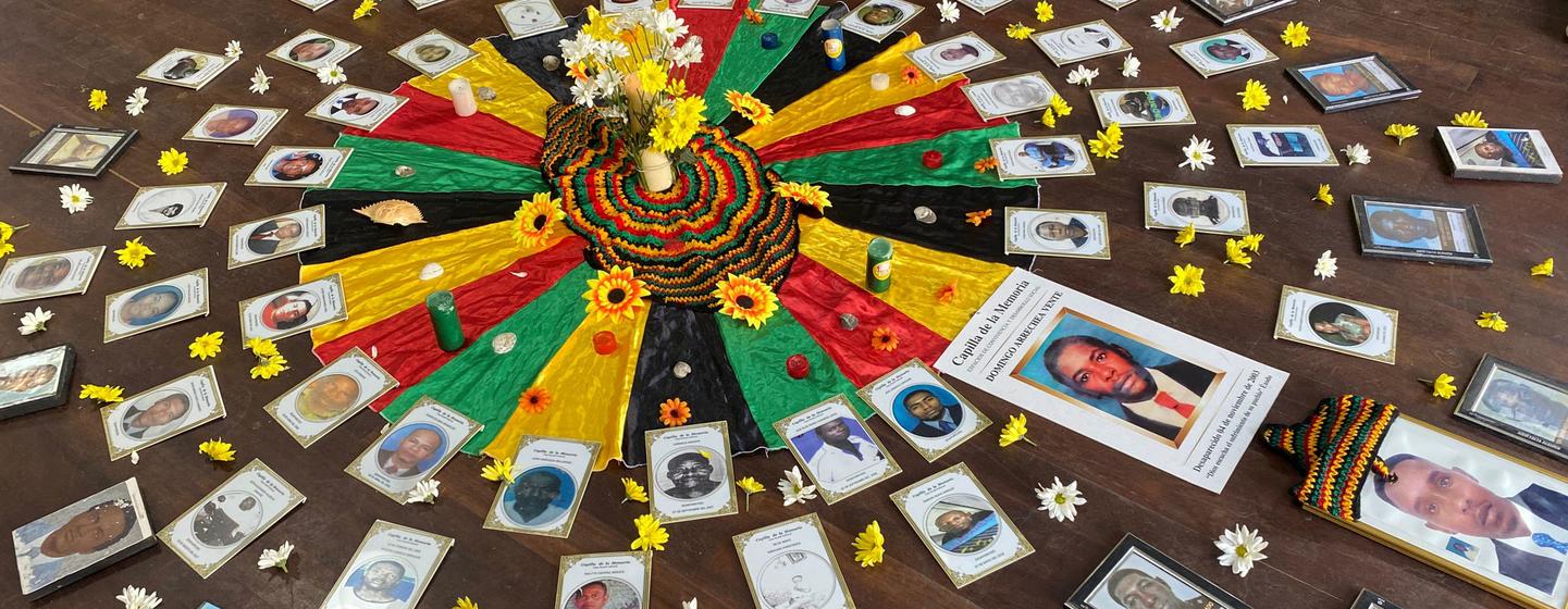 Un hommage commémoratif sur un plancher en bois présentant de nombreux portraits de personnes, un arrangement floral central et un drapeau colombien, ainsi que des fleurs jaunes et blanches.