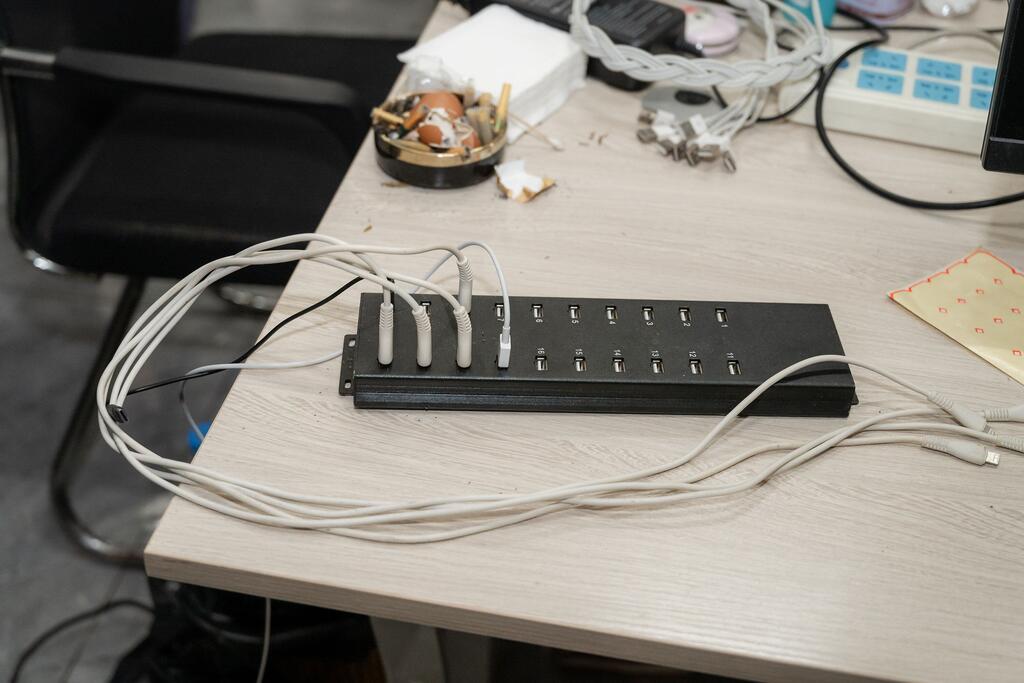 Kituo cha USB nyeusi chenye kebo nyingi nyeupe kilichounganishwa kinakaa kwenye dawati la ofisi iliyojaa fujo, likiwa limezungukwa na vifaa vingine vya kiteknolojia na takataka.