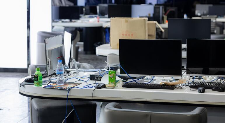 कई कंप्यूटर मॉनिटर, कीबोर्ड, और डेस्क पर पेय के साथ एक खाली कार्यालय स्थान, कंबोडिया में एक छापेमारी किए गए घोटाले केंद्र का संकेत देता है।