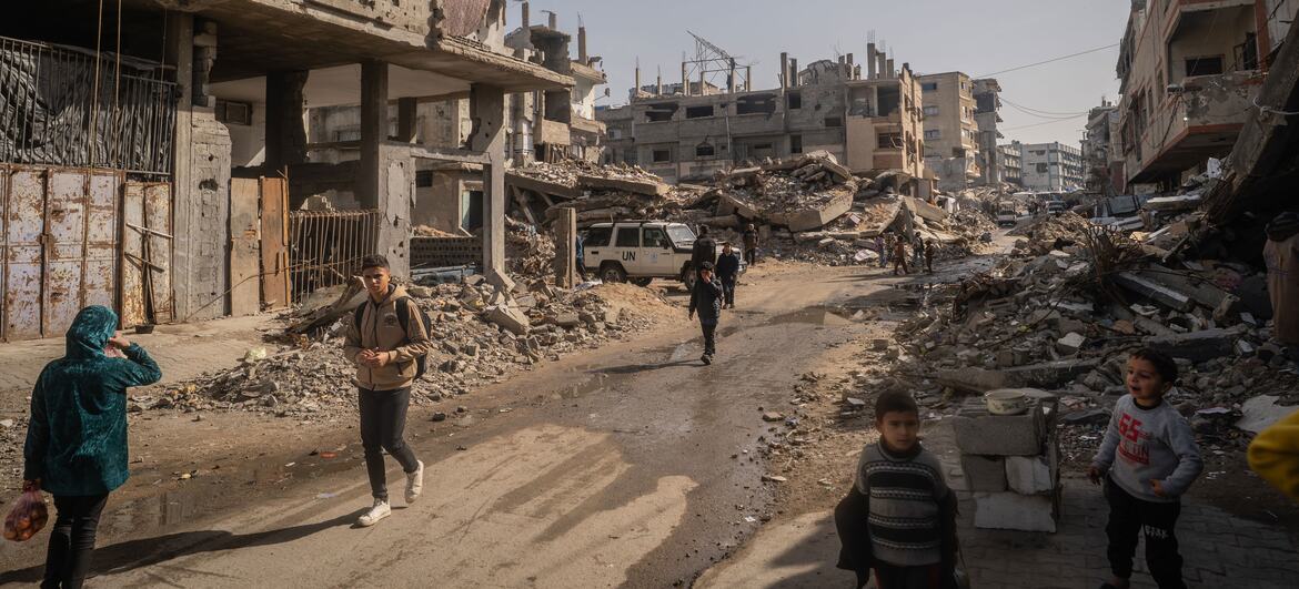 Crianças e adultos caminham por uma rua destruída em Gaza durante o Ramadã, onde as famílias se reúnem para refeições de iftar fornecidas por cozinhas comunitárias em meio a escombros e edifícios danificados.