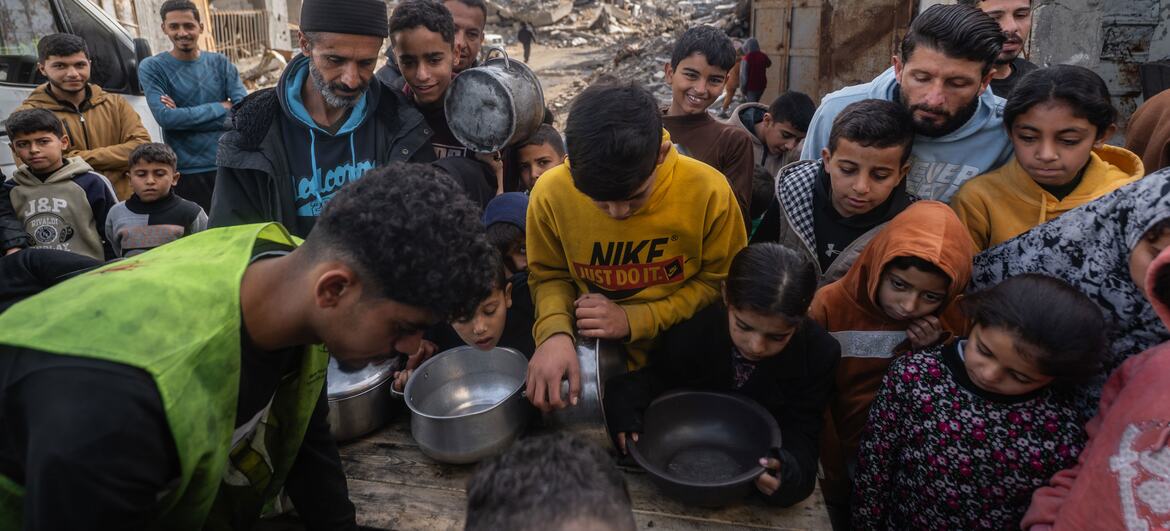 A group of Palestinian children and adults gather around a table in a war-damaged area in Gaza, receiving hot meals from a community kitchen during Ramadan.