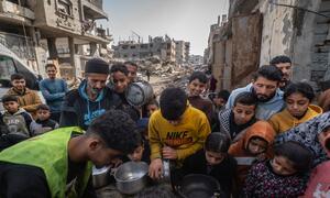 A group of Palestinian children and adults gather around a table in a war-damaged area in Gaza, receiving hot meals from a community kitchen during Ramadan.