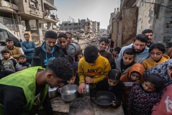 A group of Palestinian children and adults gather around a table in a war-damaged area in Gaza, receiving hot meals from a community kitchen during Ramadan.