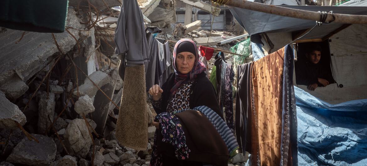 A Palestinian woman stands in a rubble-filled area, clothes hanging on a makeshift line, with damaged buildings in the background during Ramadan.
