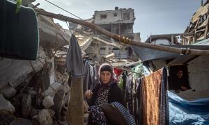 A Palestinian woman stands in a rubble-filled area, clothes hanging on a makeshift line, with damaged buildings in the background during Ramadan.