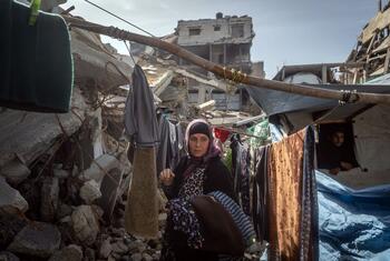 A Palestinian woman stands in a rubble-filled area, clothes hanging on a makeshift line, with damaged buildings in the background during Ramadan.
