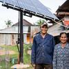 A smiling Cambodian couple standing in front of a newly installed solar panel system in their rural village, representing UNDP's work on renewable energy access and poverty eradication.