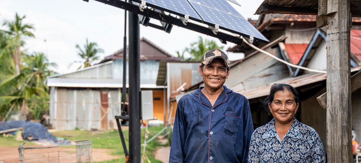 A smiling Cambodian couple standing in front of a newly installed solar panel system in their rural village, representing UNDP's work on renewable energy access and poverty eradication.