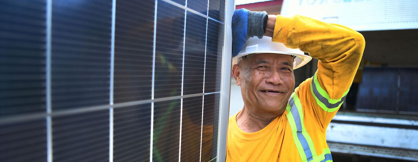 A Filipino construction worker in a yellow safety shirt and white hard hat smiles while leaning against a large solar panel during the UNDP Renewable Energy project in BARMM, Philippines.