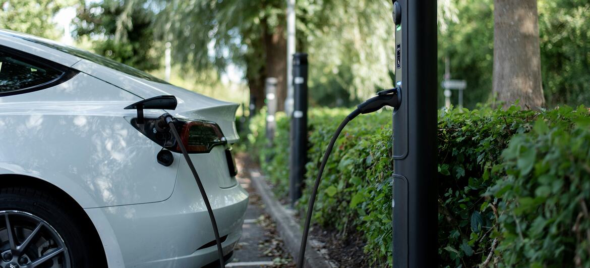 A white electric vehicle is plugged into a black charging station in a parking area surrounded by green trees and bushes.