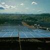 Una granja solar con filas de paneles solares en un campo verde bajo un cielo azul con nubes.