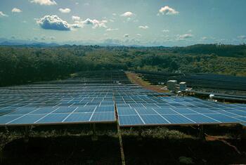 Una granja solar con filas de paneles solares en un campo verde bajo un cielo azul con nubes.