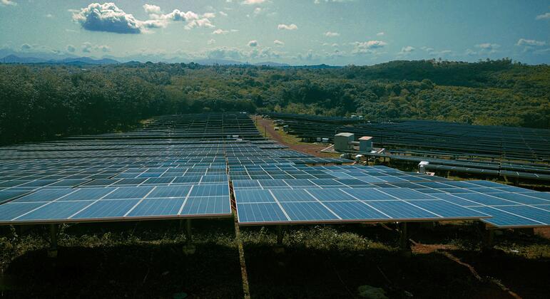 Una granja solar con filas de paneles solares en un campo verde bajo un cielo azul con nubes.