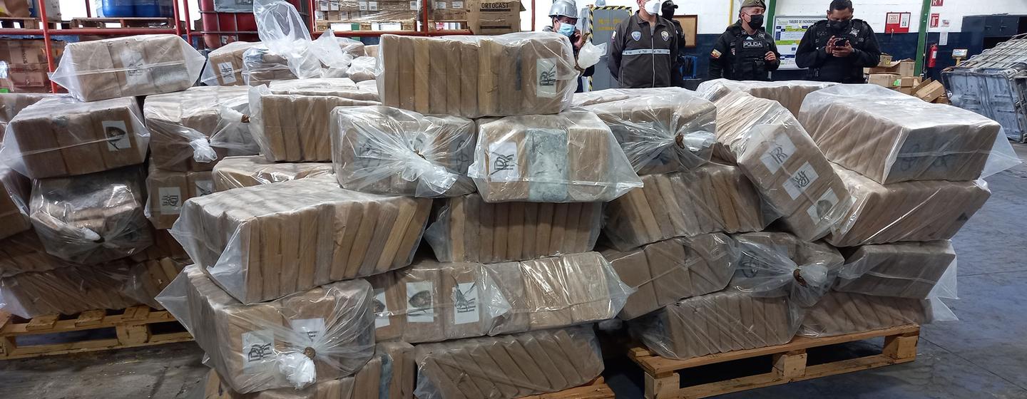 De grandes piles de paquets de cocaïne saisis enveloppés en plastique, avec des policiers observant à l'arrière-plan dans un entrepôt en Équateur.