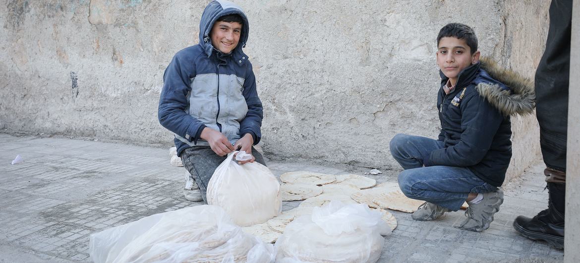 Deux garçons à Alep, en Syrie, sont assis sur un trottoir à côté de sacs et de piles de pain plat lors d'une mission de distribution alimentaire du gouvernement américain Fletcher menée par OCHA.