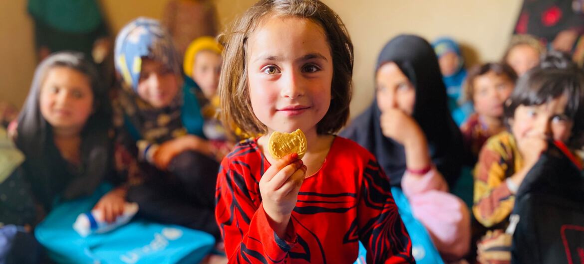 A young girl in Afghanistan holds a fortified biscuit while other children sit around her with blue backpacks.