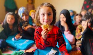 A young girl in Afghanistan holds a fortified biscuit while other children sit around her with blue backpacks.