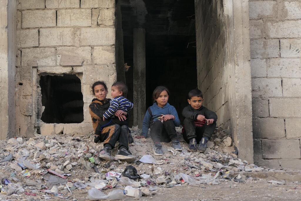Quatre enfants, dont un tenu par une fillette plus âgée, sont assis au milieu des décombres d'un bâtiment en ruines.