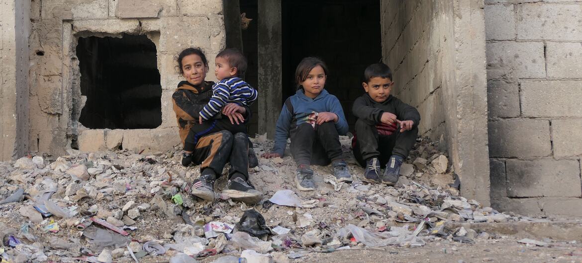 Four children, including one being held by an older girl, sit amidst rubble in a ruined building.