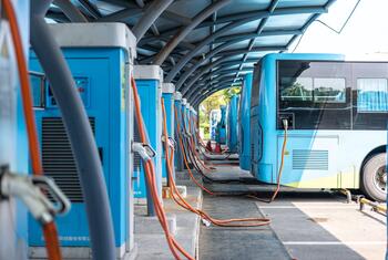 A row of blue electric buses parked at a charging station, connected by thick orange cables.