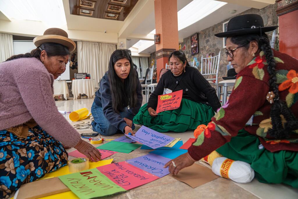 Cuatro mujeres vestidas con trajes tradicionales peruanos participan en un taller, trabajando con papel de colores para crear un mural en el suelo.