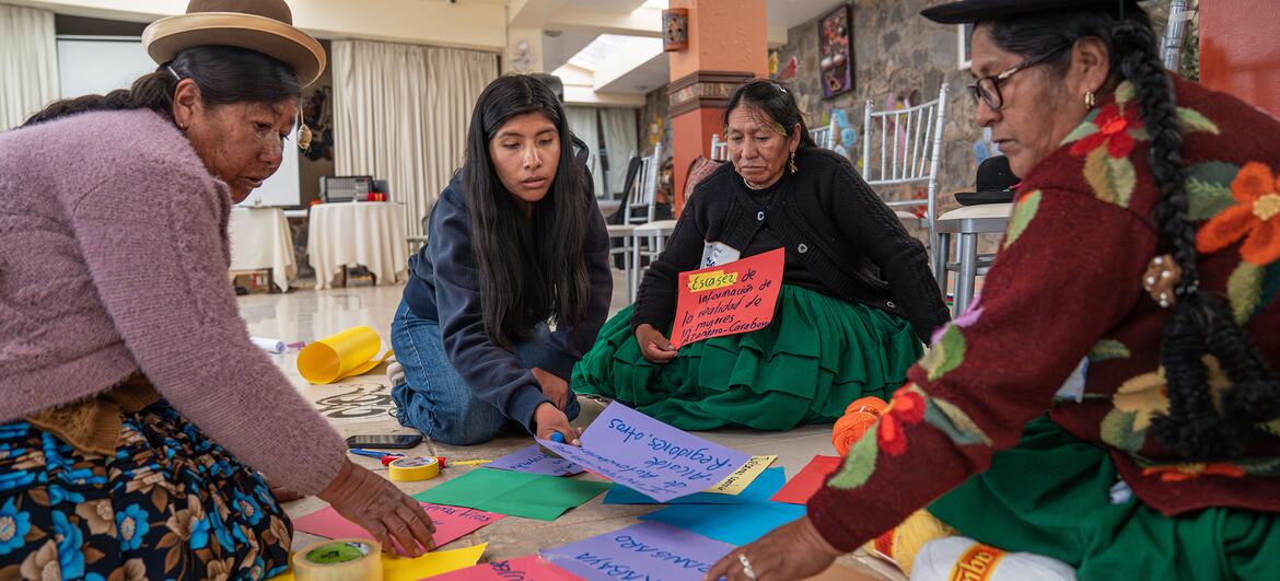 Four women in traditional Peruvian attire are participating in a workshop, working with colored paper to create a mural on the floor.