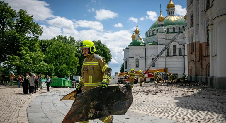 Un pompier ukrainien transporte un gros morceau de gravats devant la Laure des Grottes de Kyiv, tandis que d'autres secouristes et un camion de pompiers sont visibles en arrière-plan.